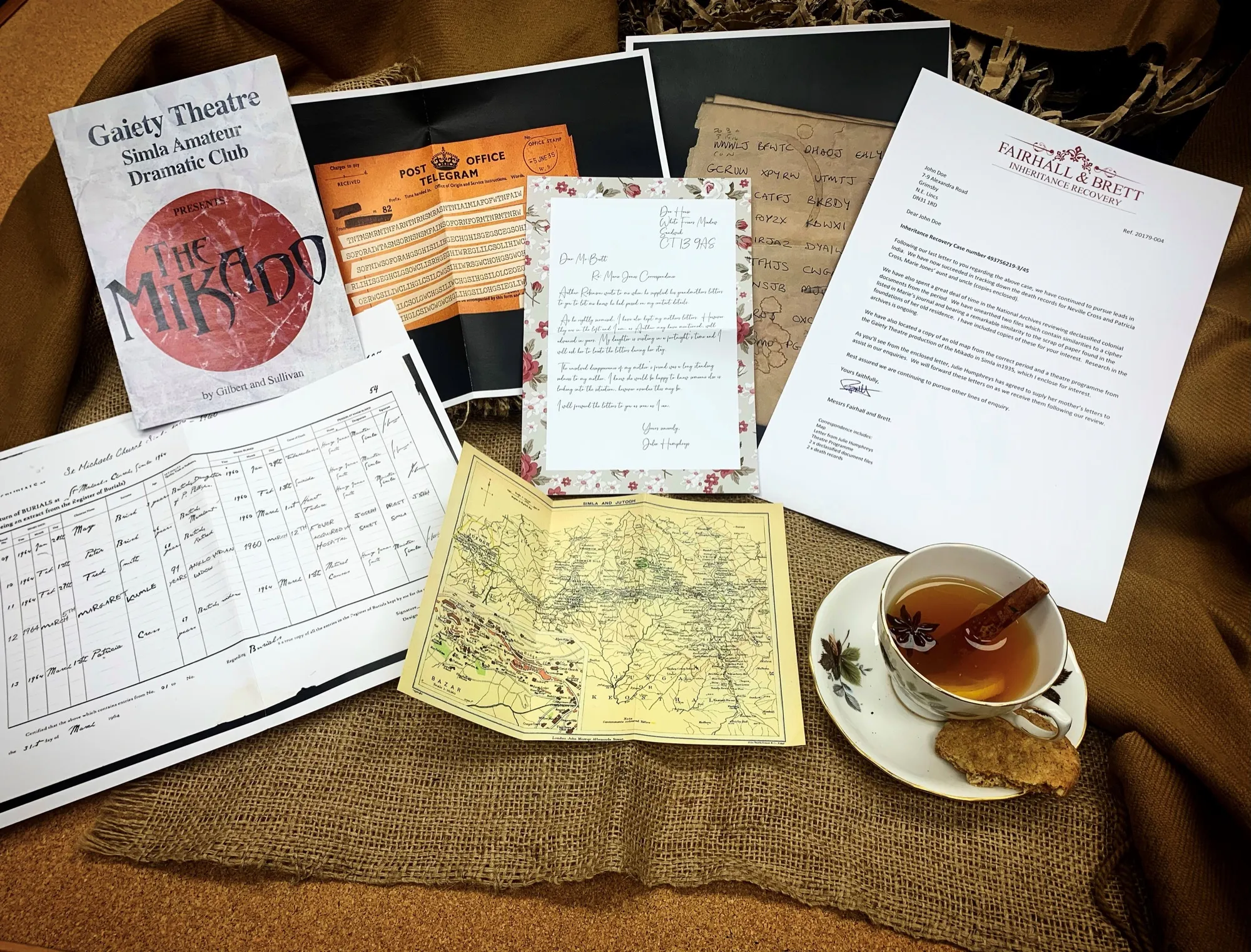 The Inheritance of Murder case file spread across hessian — a Gaiety Theatre Simla programme for The Mikado, an orange Post Office telegram, a hand-coloured map of the Simla hills, a floral handwritten letter and the Fairhall & Brett inheritance-recovery letter, beside a cup of spiced tea